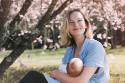 Eine junge Frau sitzt im Park vor einem blühendem Baum mit einem Baby im Arm. 